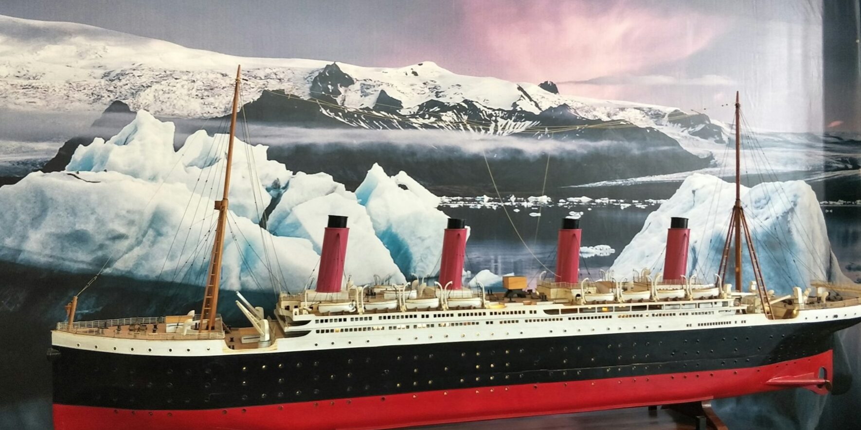 Schaalmodel van de Titanic met vier rode schoorstenen, geplaatst voor een achtergrond van ijsbergen en besneeuwde bergen.