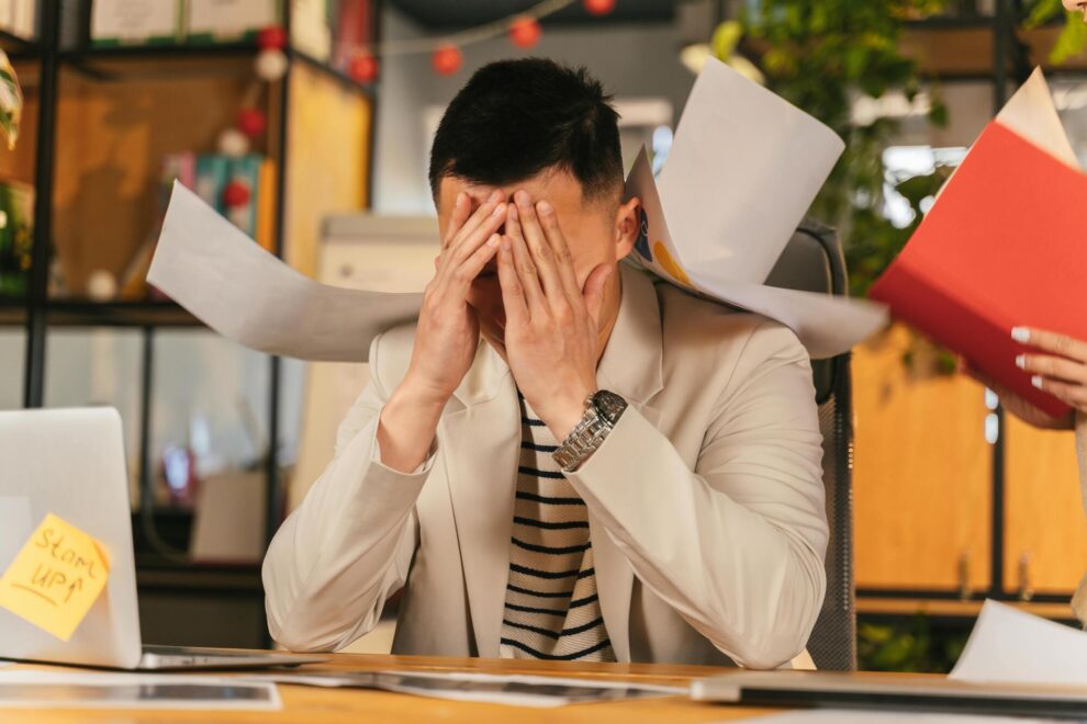 Man zit gestrest aan bureau terwijl papieren om hem heen vliegen op kantoor Man zit gestrest aan bureau terwijl papieren om hem heen vliegen op kantoor