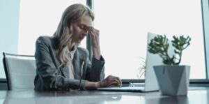 Vrouw zit vermoeid achter laptop en houdt hand tegen voorhoofd op kantoor